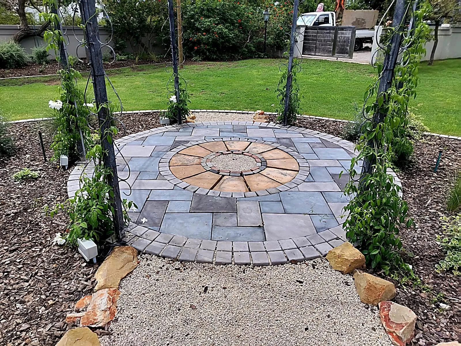 A round paved feature with a metal gazebo, planted beds and a stone path in a Jeffreys Bay garden